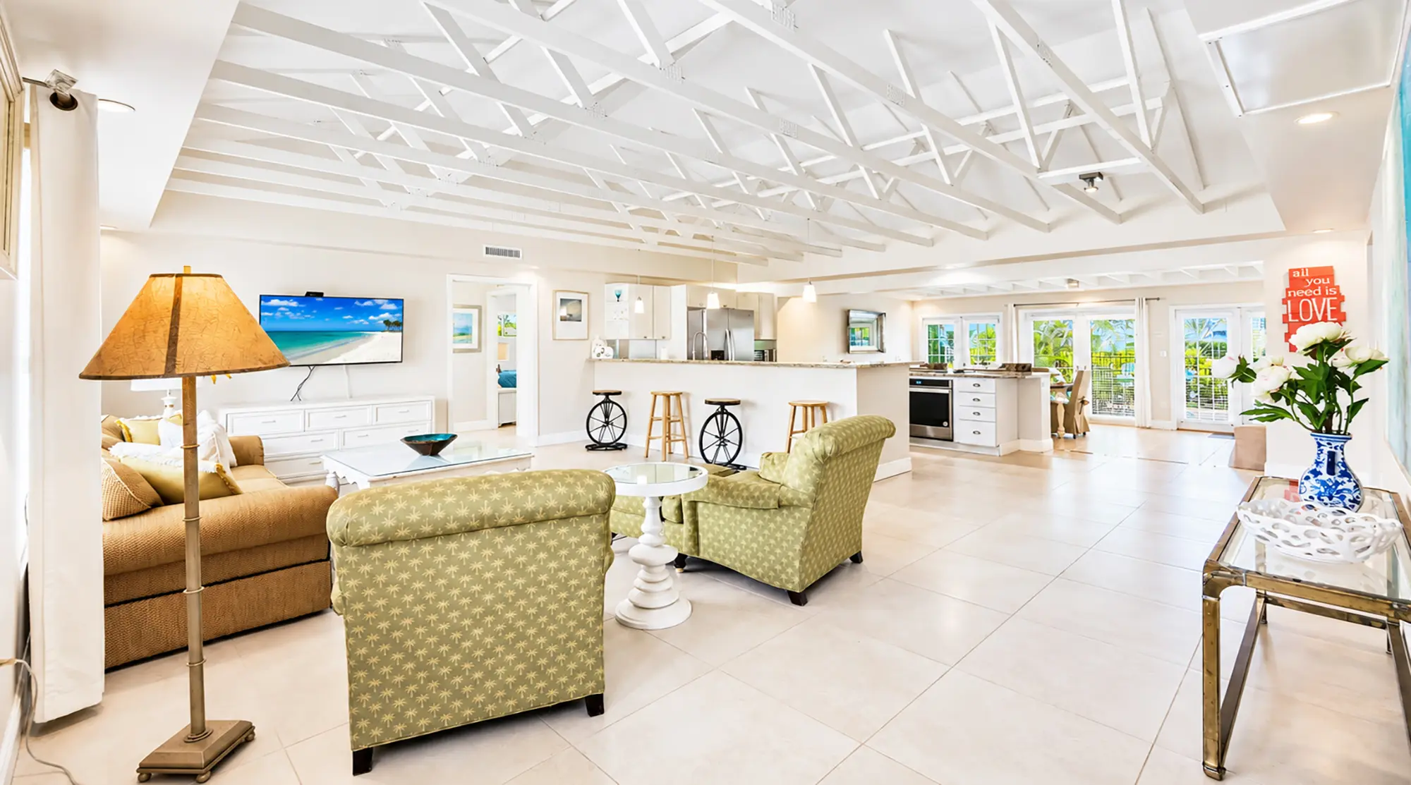 Open-concept living room and kitchen with vaulted ceilings, beige sofas, green armchairs, a central breakfast bar with bicycle-wheel stools, and tile flooring.