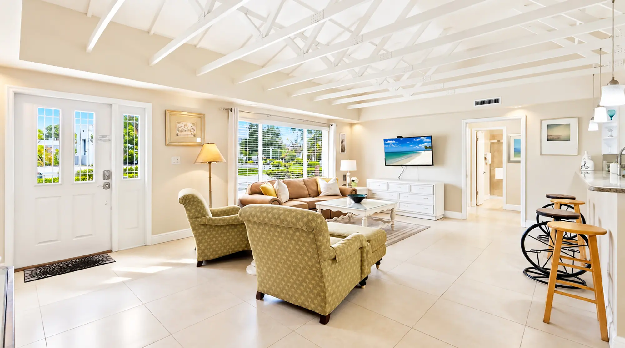 Living room with two patterned armchairs, a tan sofa, a television mounted on the wall, and a kitchen island with bar stools.