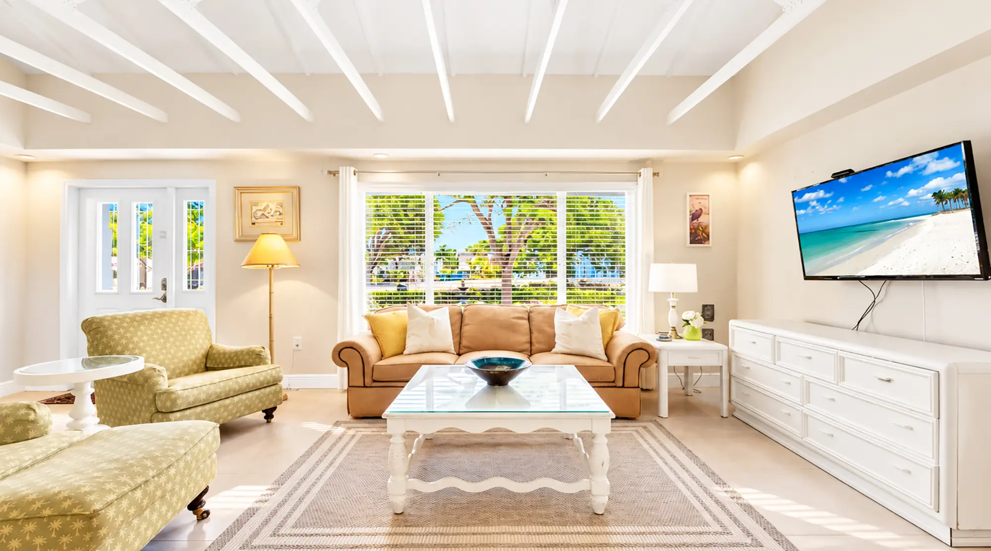 A living room in a Naples waterfront vacation rental featuring a beige sofa, two patterned armchairs, a white coffee table, and a television mounted on the wall displaying a beach scene.