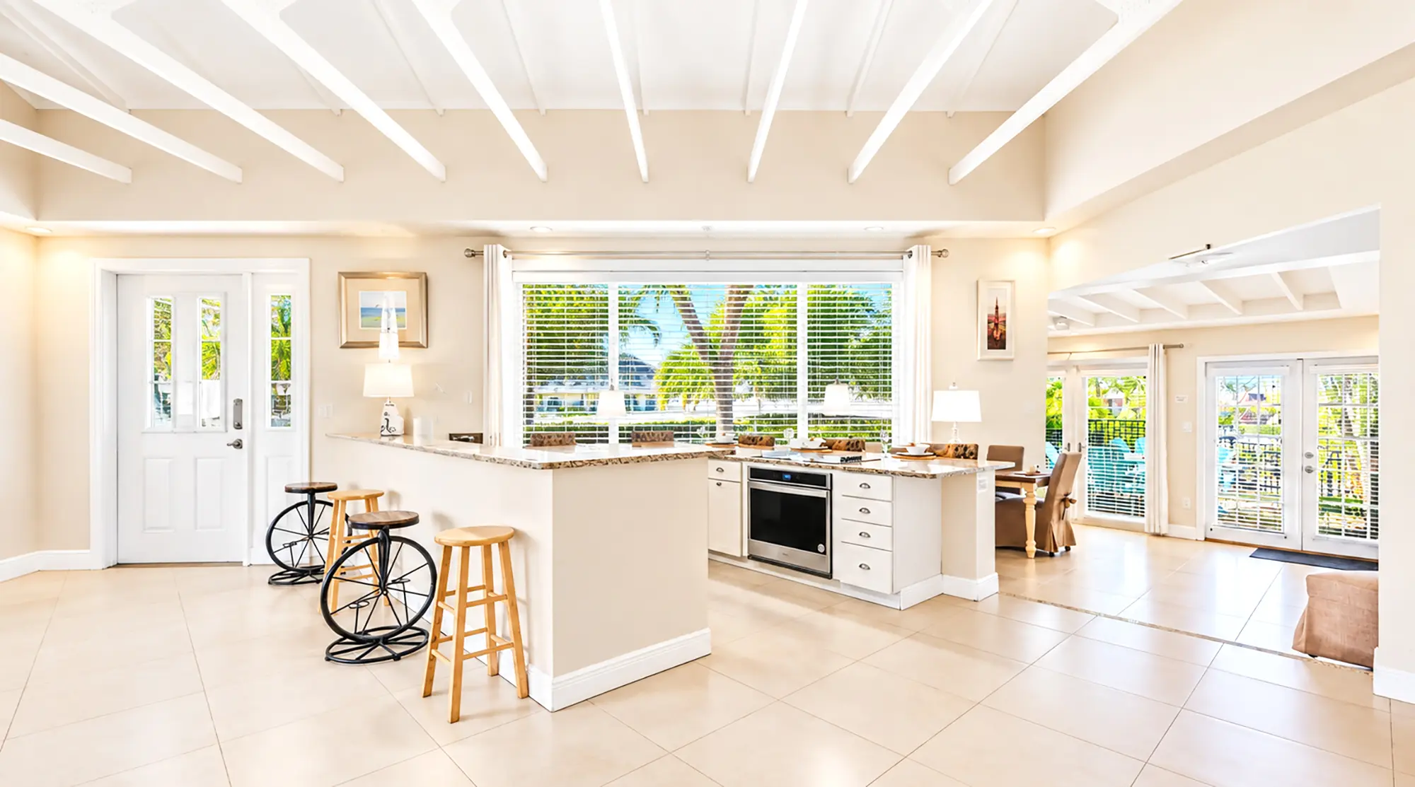 Kitchen island with a countertop, two bicycle-style stools, and a built-in oven, overlooking a view of palm trees through large windows.