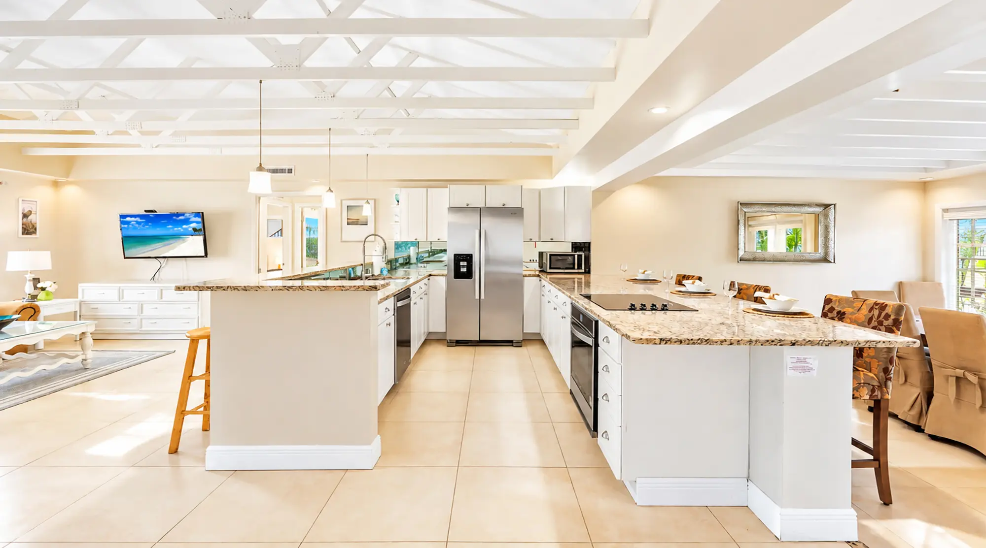 A spacious kitchen with light-colored cabinets, stainless steel appliances, and a granite-topped island, opening into a living room with a television mounted on the wall.