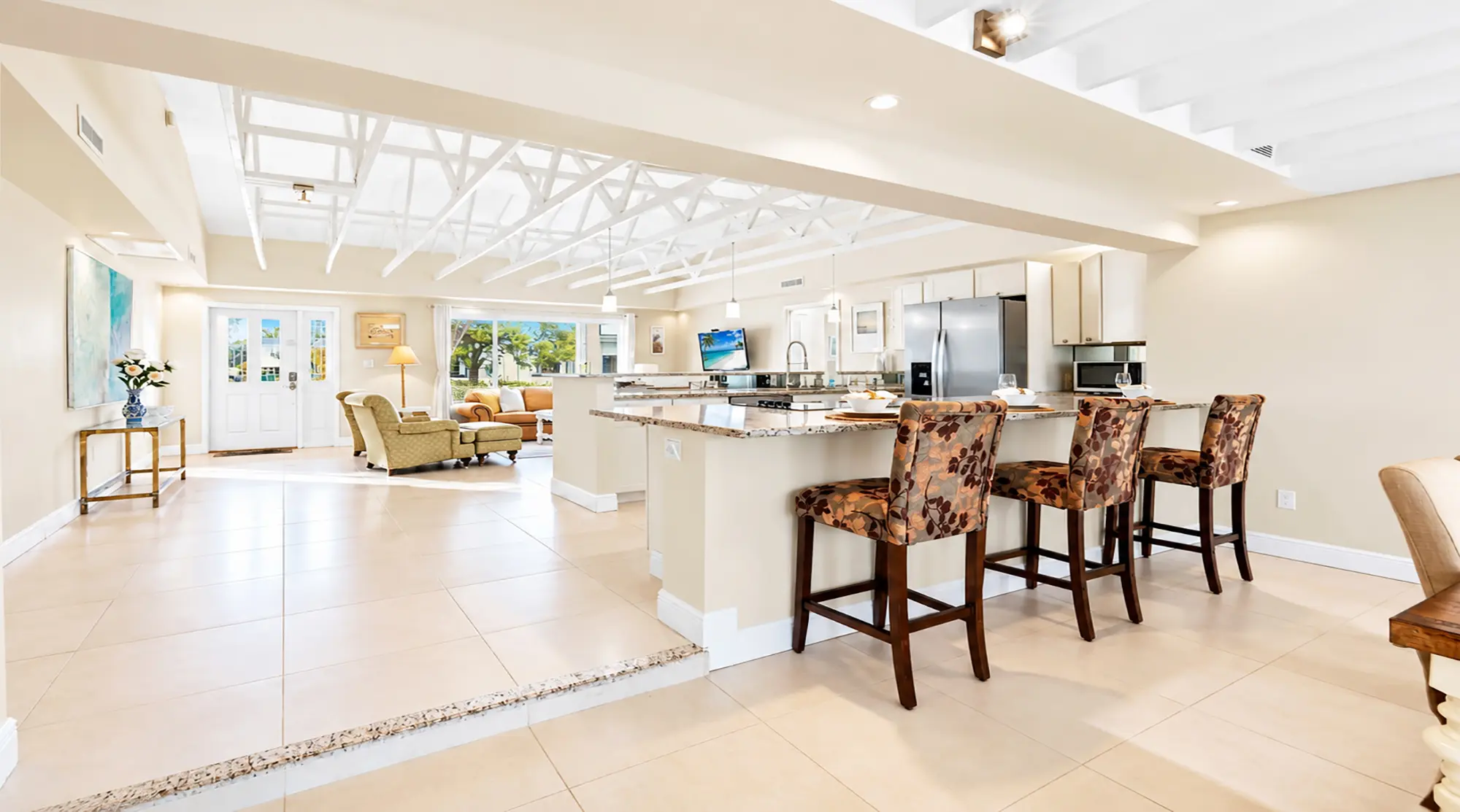 Open-concept kitchen and living area with a stone-topped island, floral-patterned bar stools, and a view into a sunlit living room with vaulted white ceiling beams.