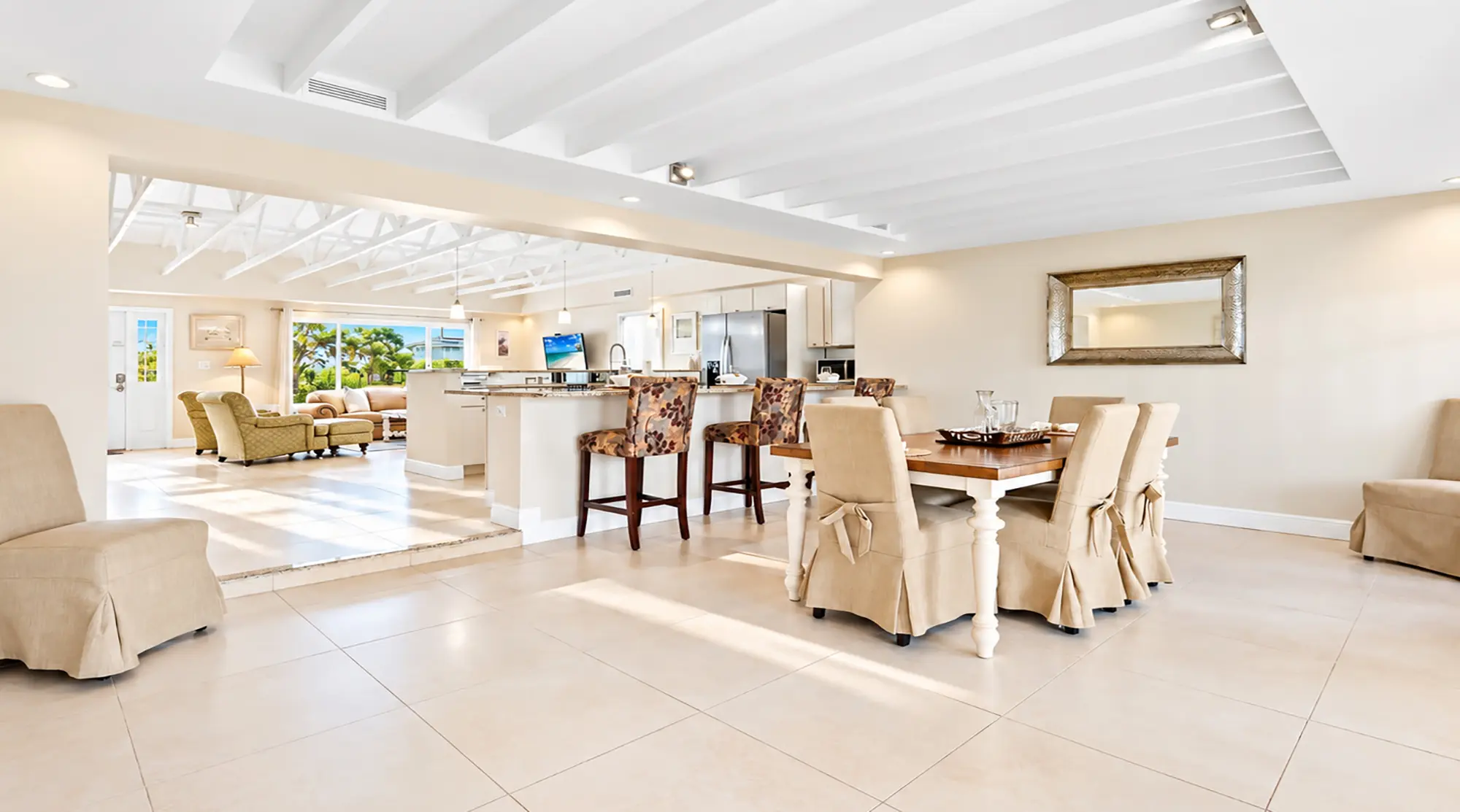 An open-concept living area featuring a dining table with slipcovered chairs in the foreground, a kitchen island with bar stools, and a sunlit living room in the back.