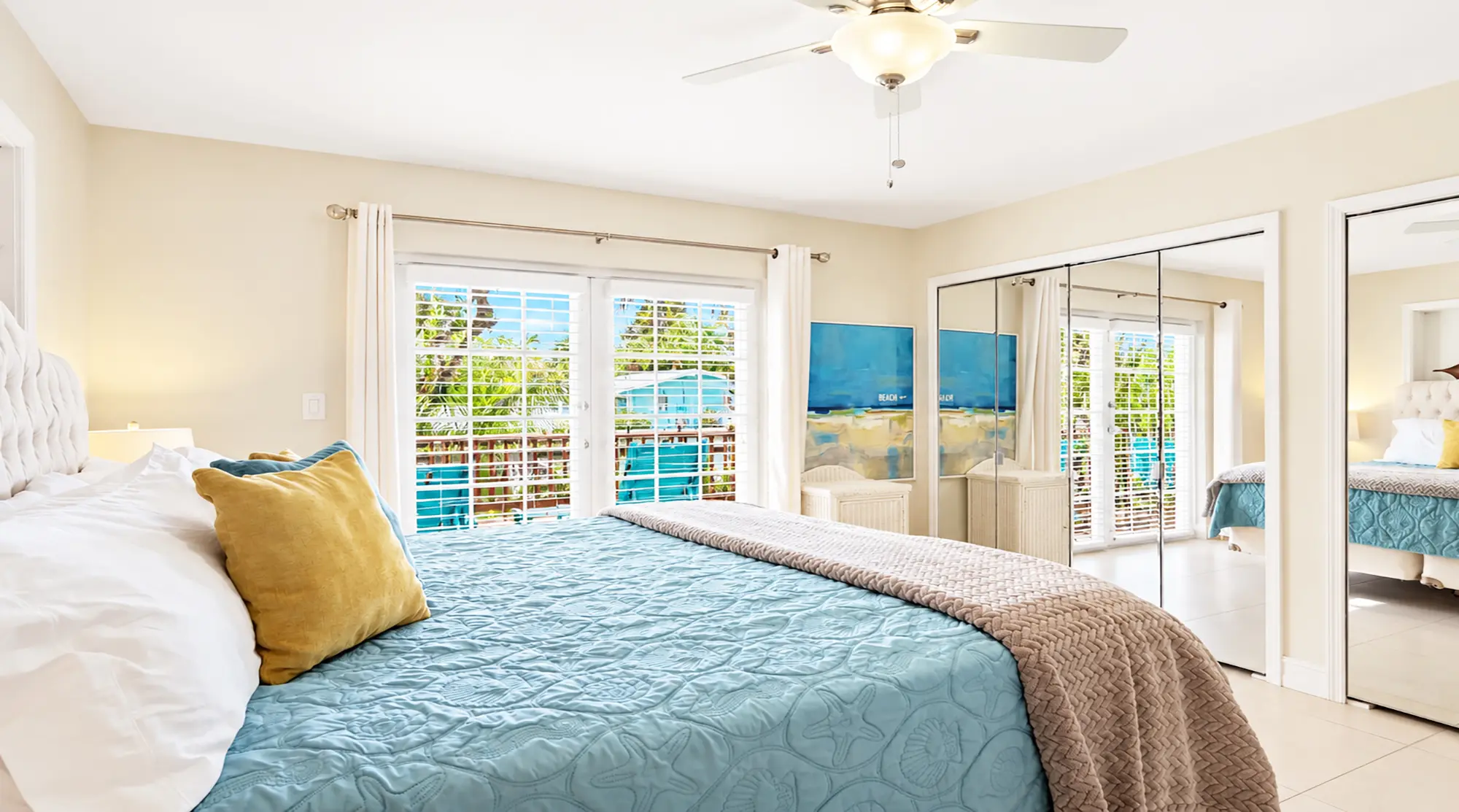 Bedroom with a bed dressed in a blue quilt and yellow pillow, featuring glass sliding doors leading to a balcony and large mirrored closet doors.