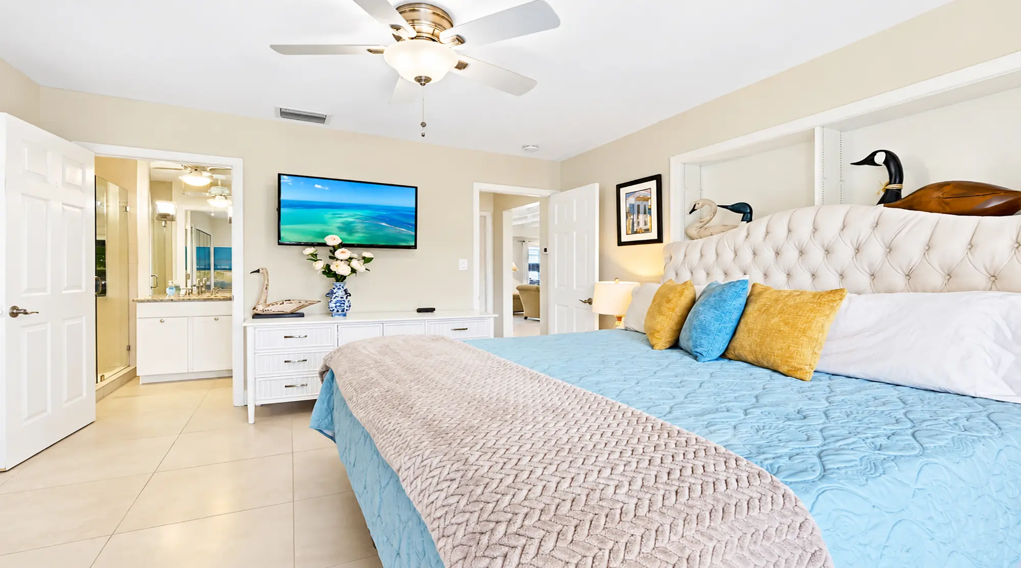 Bedroom with a bed featuring a tufted headboard, blue quilt, and yellow pillows, a mounted television, and a doorway leading to a bathroom.
