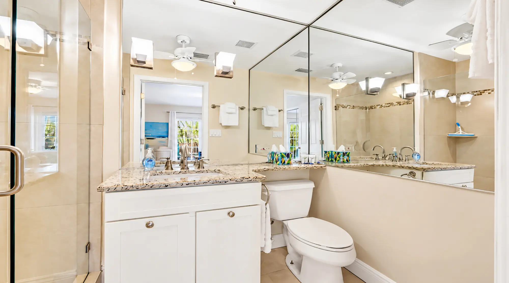 A bathroom with a white vanity, a toilet, and a large wall mirror reflecting a view into an adjacent bedroom.