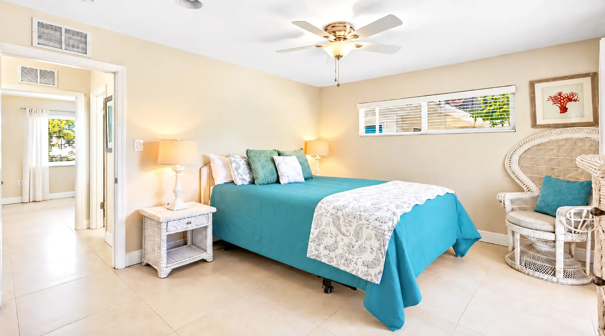A bedroom with a turquoise bed, a white wicker nightstand, a matching wicker chair with a turquoise pillow, and a ceiling fan.