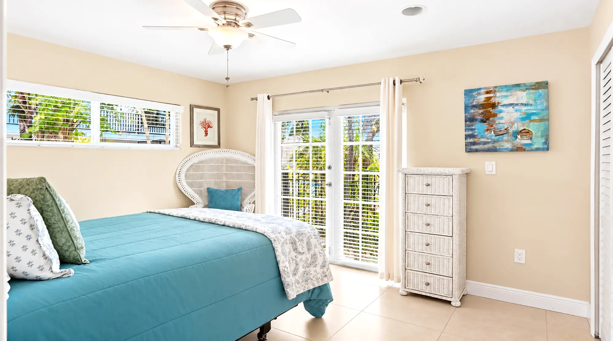 Bedroom with a turquoise bedspread, white wicker chair, tall white dresser, and glass doors leading to a balcony.