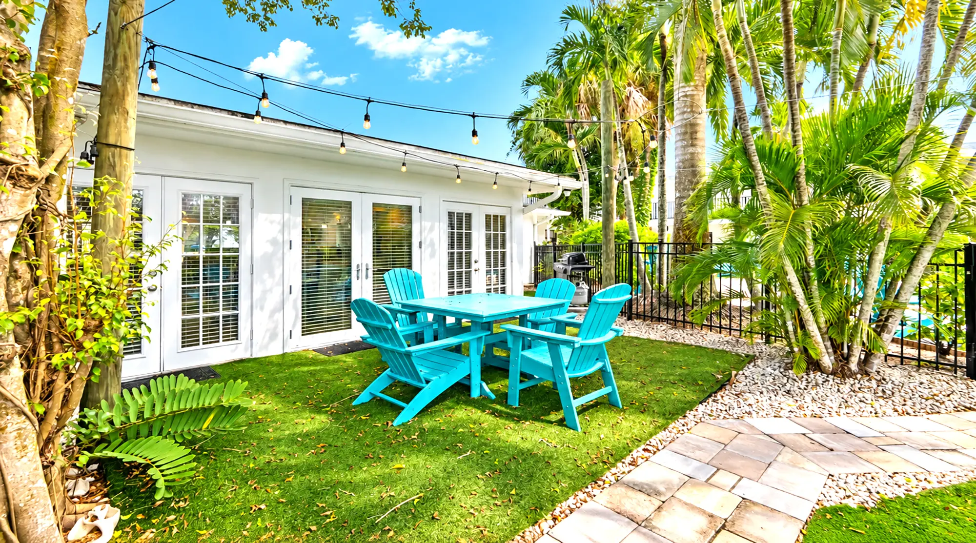 Turquoise Adirondack chairs and a matching table on a patch of green lawn outside a white house with string lights.