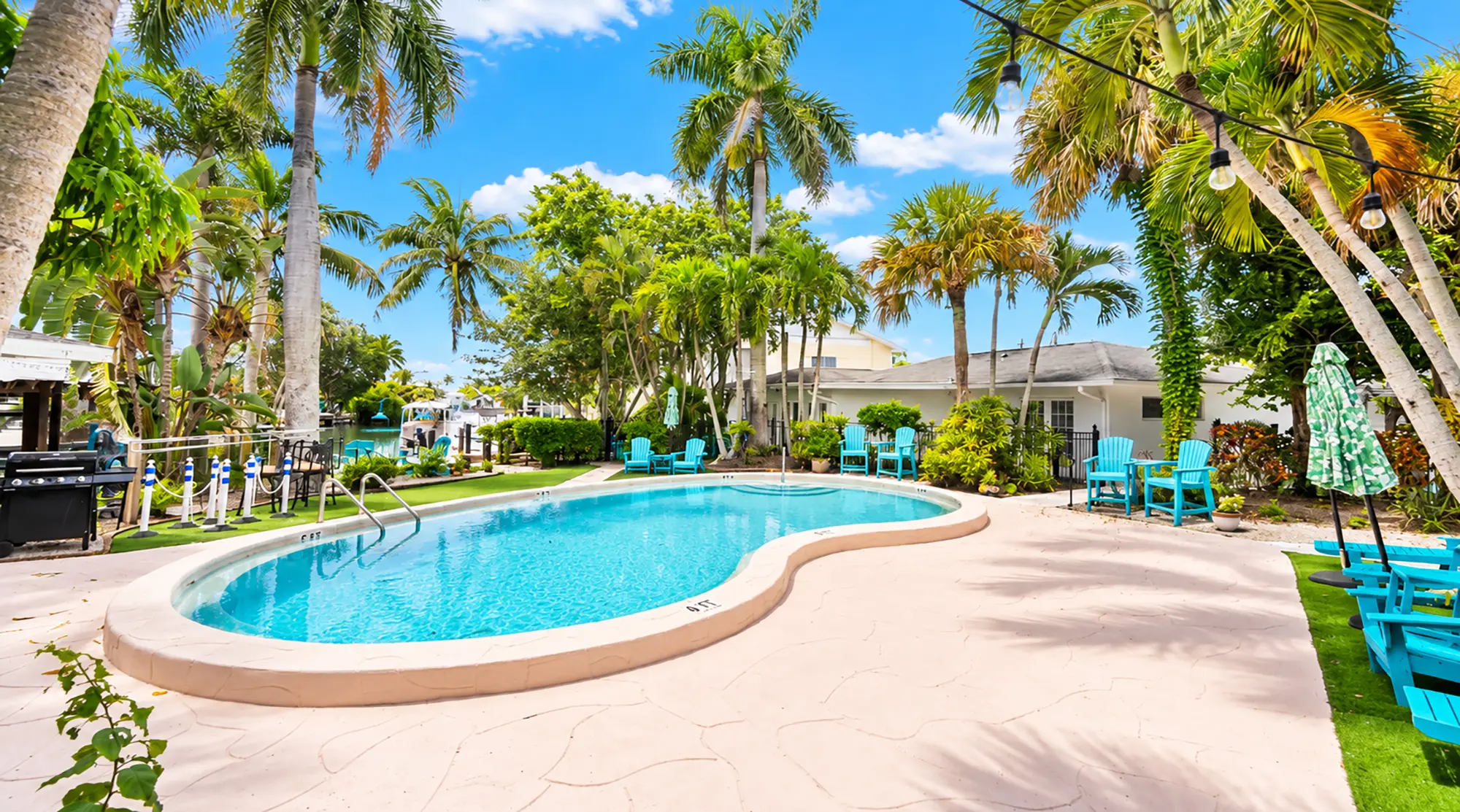 A kidney-shaped swimming pool on a patio surrounded by palm trees and bright blue Adirondack chairs.