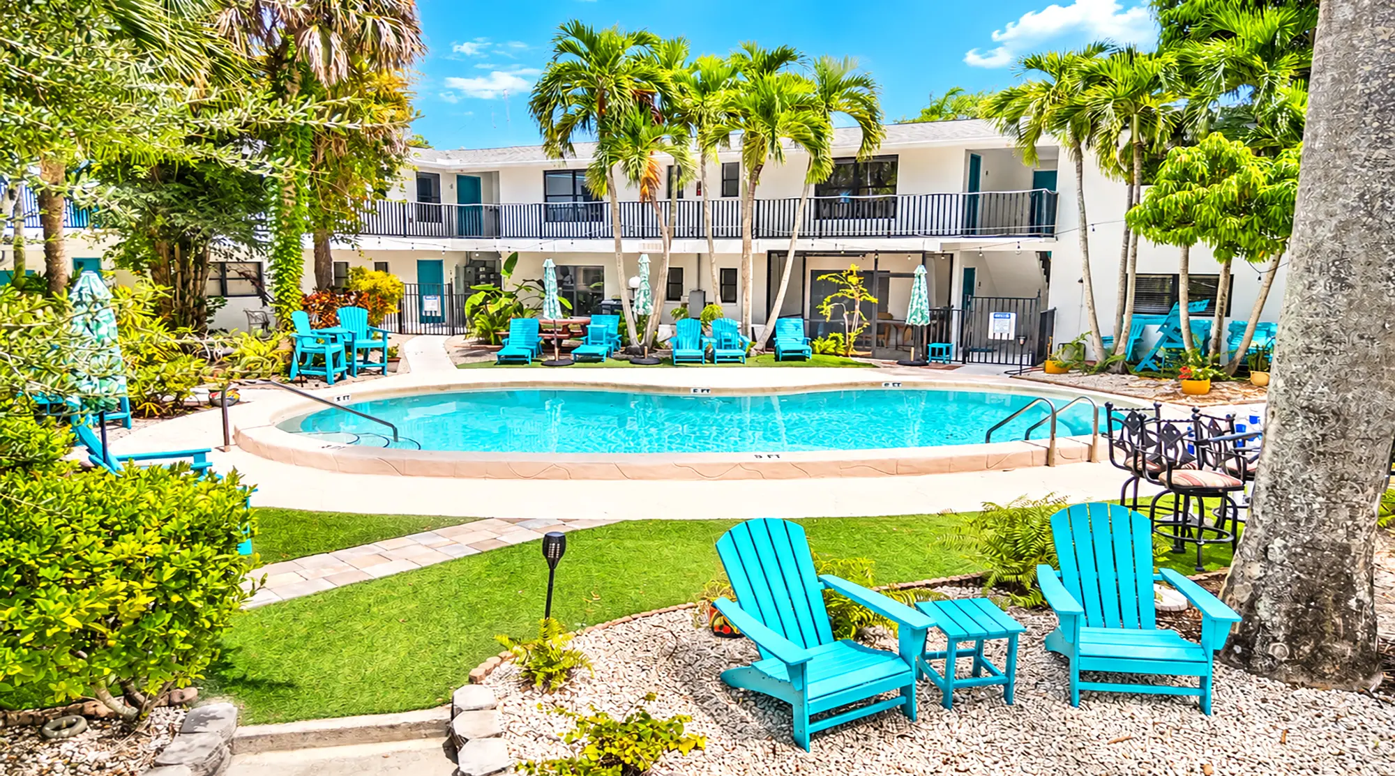 A courtyard with a swimming pool surrounded by palm trees, two-story apartment buildings, and bright blue lounge chairs.