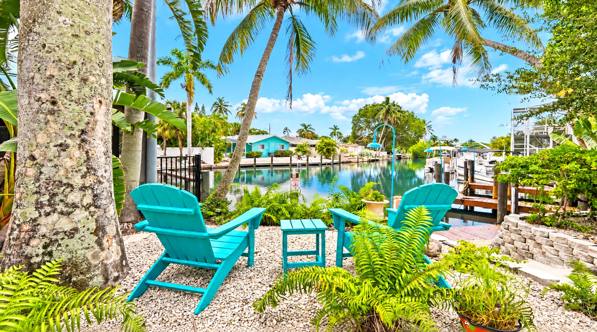 Two teal Adirondack chairs and a matching side table on a gravel patio overlooking a canal with docked boats.