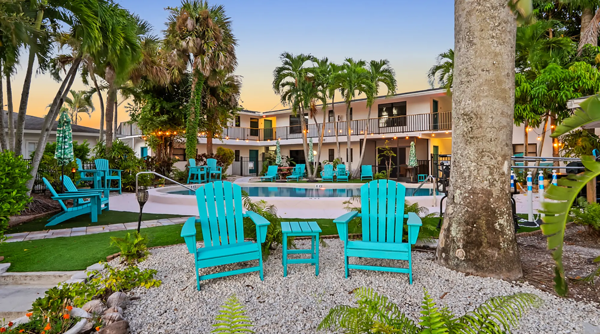 Two turquoise Adirondack chairs and a small matching side table sit on a gravel patio facing a hotel swimming pool surrounded by palm trees.